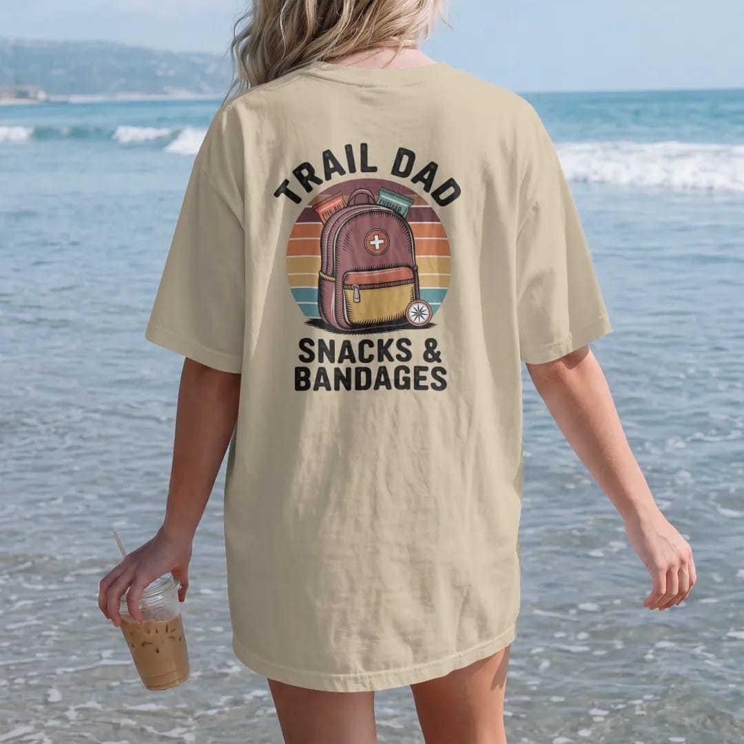 Woman on the beach in a Sandstone tee, back view: a fully loaded daypack with a first-aid patch, snacks peeking out and a small compass, faded 70s souvenir style, lettered "TRAIL DAD / SNACKS & BANDAGES" in chunky rounded 70s letters with a drop…