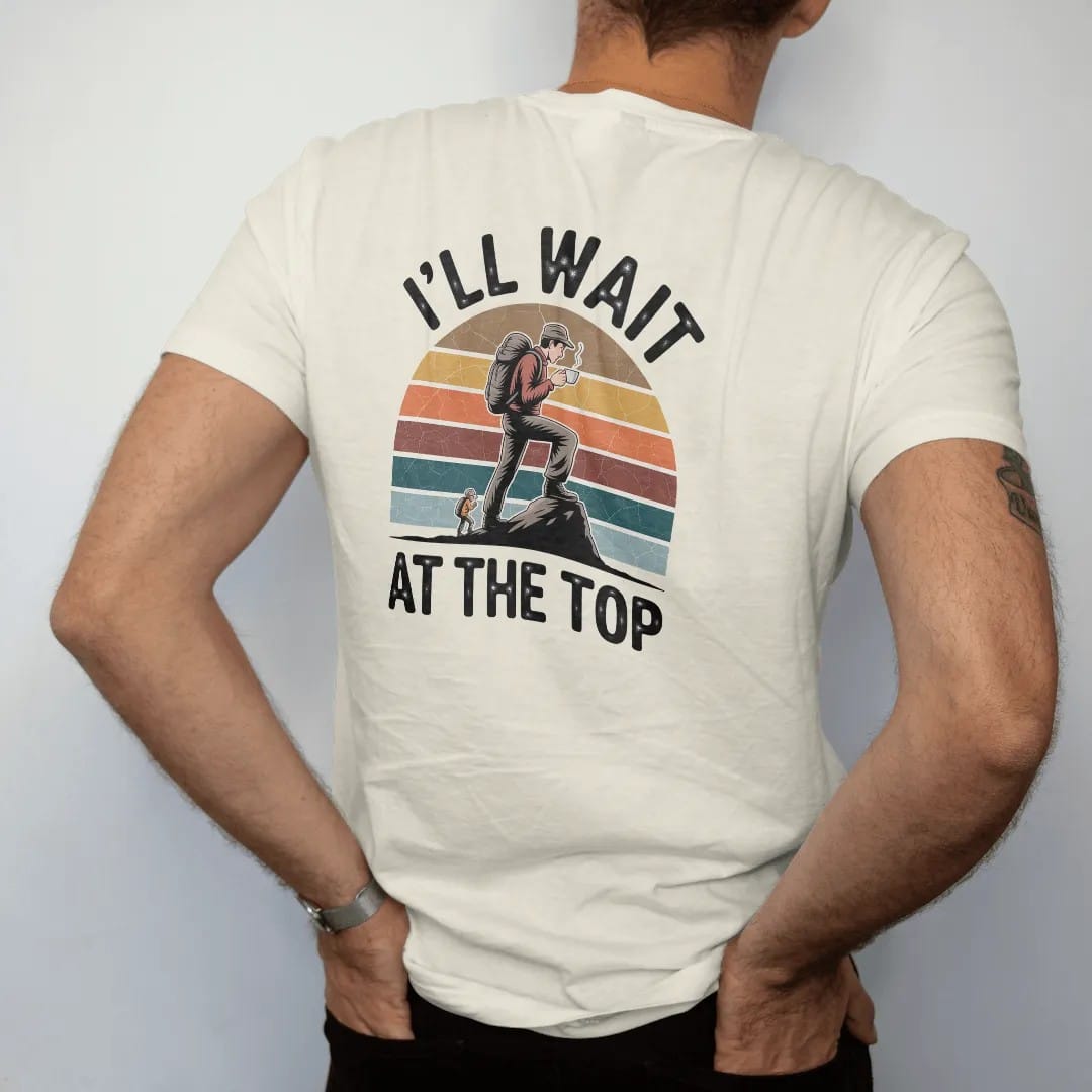 Man wearing a Ivory tee, seen from the back: a smug hiker on a summit rock sipping coffee while a tiny friend climbs below, faded 70s souvenir style, lettered "I'LL WAIT / AT THE TOP" in chunky rounded 70s letters with a drop shadow and a retro…