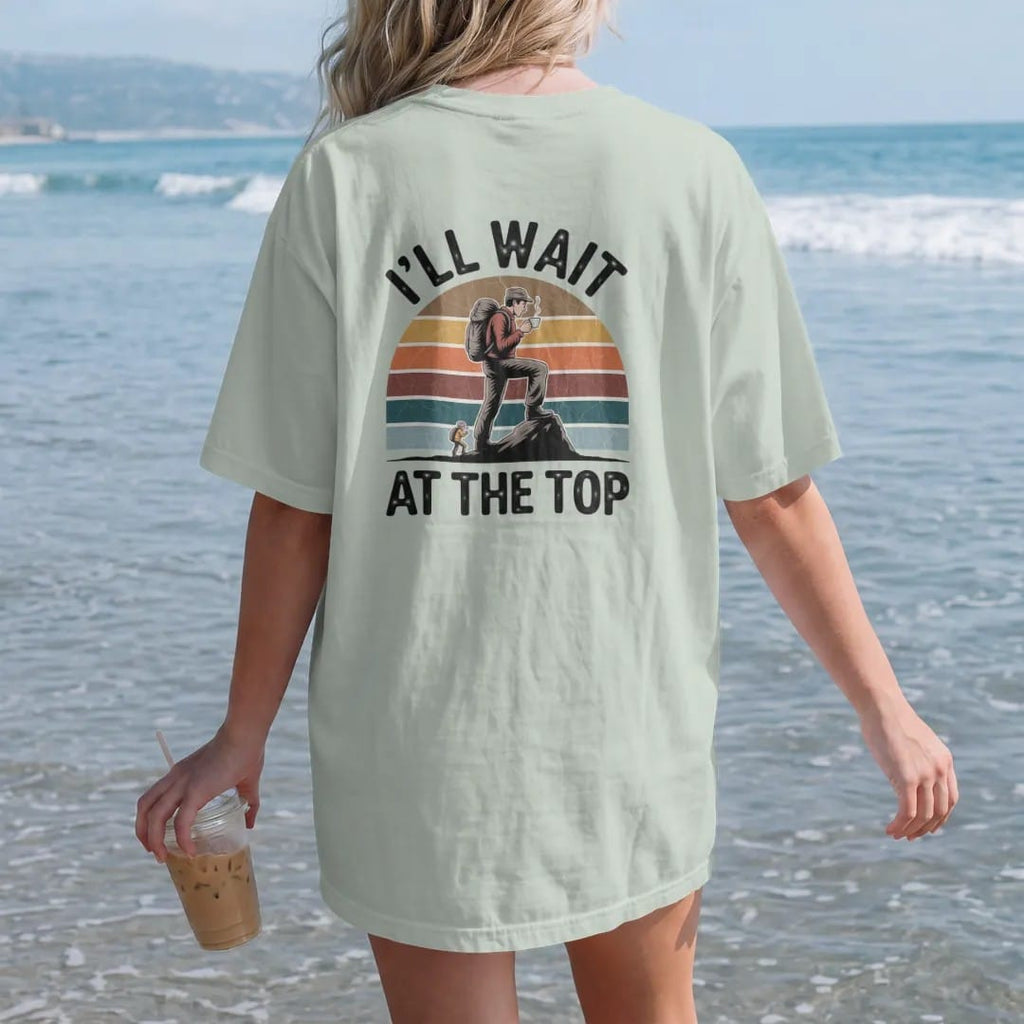Woman on the beach in a Bay tee, back view: a smug hiker on a summit rock sipping coffee while a tiny friend climbs below, faded 70s souvenir style, lettered "I'LL WAIT / AT THE TOP" in chunky rounded 70s letters with a drop shadow and a retro…