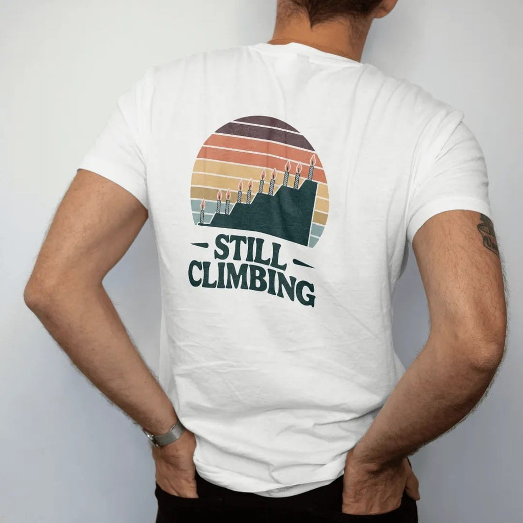 Man wearing a White tee, seen from the back: a long elevation profile line rising steadily with small birthday candles as summit flags, faded 70s souvenir style, lettered "STILL CLIMBING" in chunky rounded 70s letters with a drop shadow, printed in…
