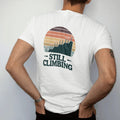 Man wearing a White tee, seen from the back: a long elevation profile line rising steadily with small birthday candles as summit flags, faded 70s souvenir style, lettered "STILL CLIMBING" in chunky rounded 70s letters with a drop shadow, printed in…