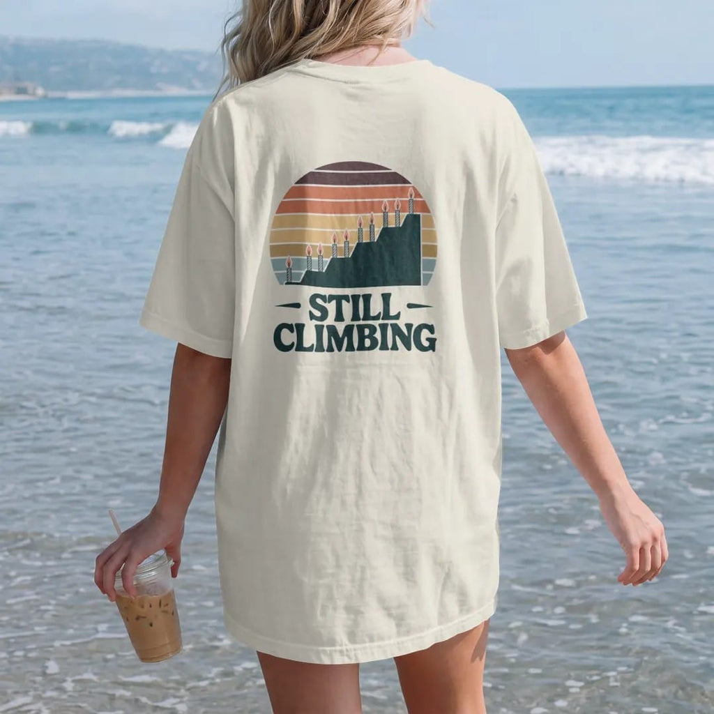 Woman on the beach in a Ivory tee, back view: a long elevation profile line rising steadily with small birthday candles as summit flags, faded 70s souvenir style, lettered "STILL CLIMBING" in chunky rounded 70s letters with a drop shadow, printed…