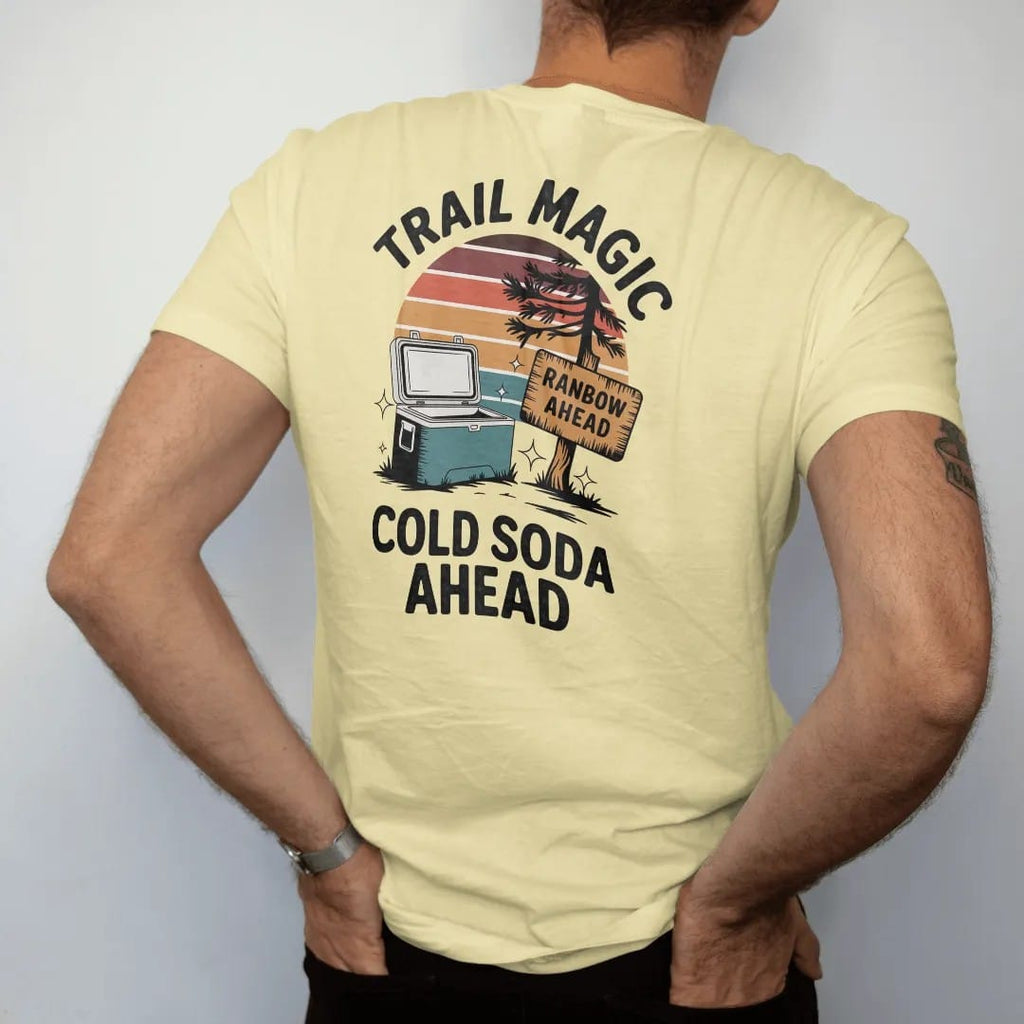 Man wearing a Butter tee, seen from the back: a cooler sitting by a trailside tree with sparkles around it and a hand-painted cardboard sign, faded 70s souvenir style, lettered "TRAIL MAGIC / COLD SODA AHEAD" in chunky rounded 70s letters with a…