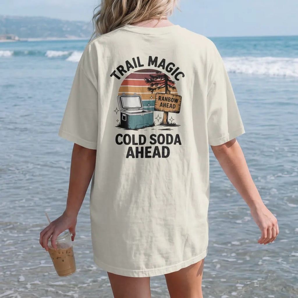 Woman on the beach in a Ivory tee, back view: a cooler sitting by a trailside tree with sparkles around it and a hand-painted cardboard sign, faded 70s souvenir style, lettered "TRAIL MAGIC / COLD SODA AHEAD" in chunky rounded 70s letters with a…