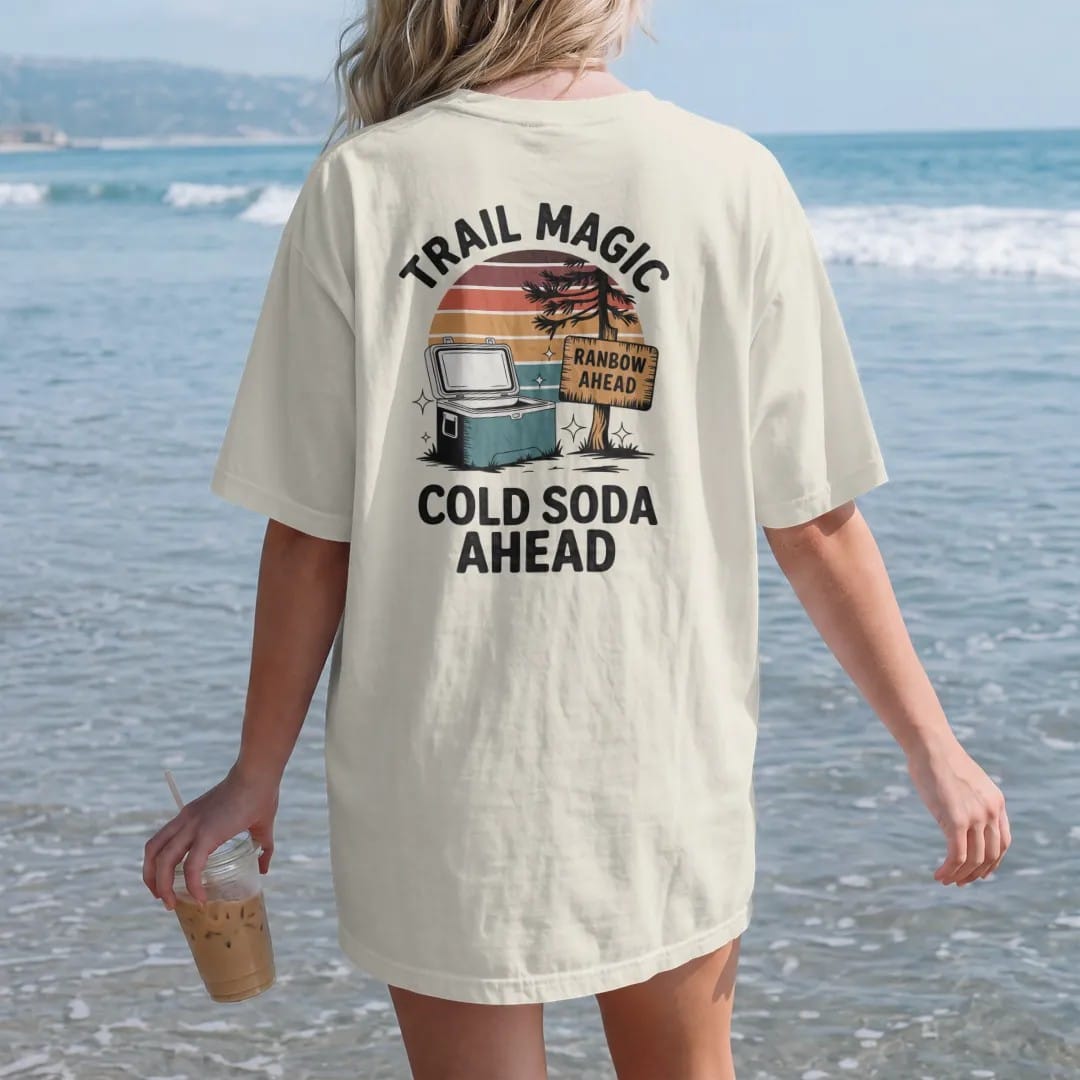 Woman on the beach in a Ivory tee, back view: a cooler sitting by a trailside tree with sparkles around it and a hand-painted cardboard sign, faded 70s souvenir style, lettered "TRAIL MAGIC / COLD SODA AHEAD" in chunky rounded 70s letters with a…