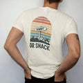 Man wearing a Ivory tee, seen from the back: a trail junction sign with one arrow pointing to a peak and one to a picnic table, faded 70s souvenir style, lettered "SUMMIT / OR SNACK" in chunky rounded 70s letters with a drop shadow, printed in…