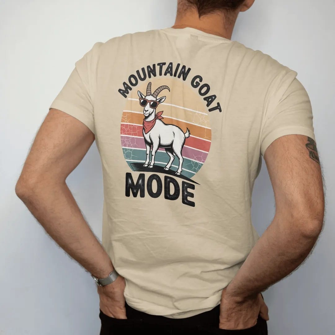 Man wearing a Sandstone tee, seen from the back: a confident mountain goat standing on a cliff edge with tiny sunglasses and a bandana, faded 70s souvenir style, lettered "MOUNTAIN GOAT / MODE" in chunky rounded 70s letters with a drop shadow,…