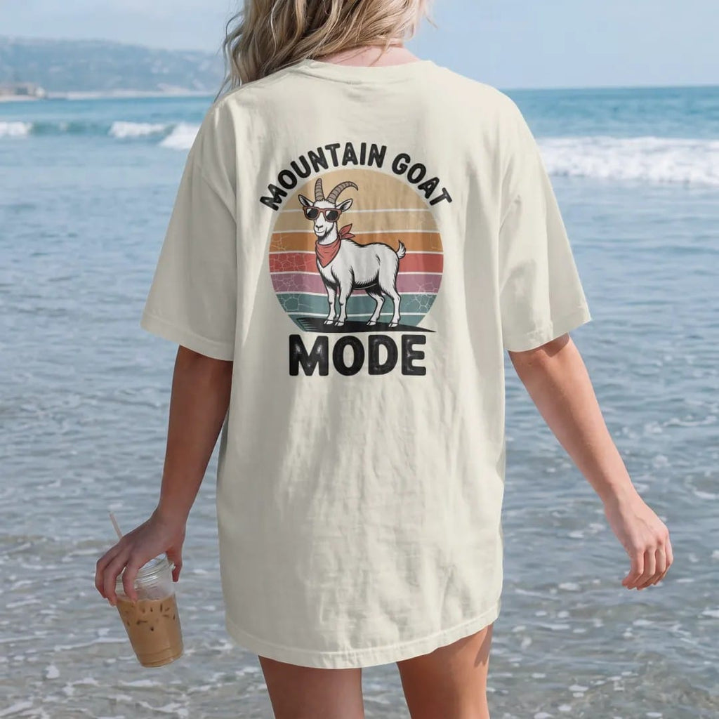 Woman on the beach in a Ivory tee, back view: a confident mountain goat standing on a cliff edge with tiny sunglasses and a bandana, faded 70s souvenir style, lettered "MOUNTAIN GOAT / MODE" in chunky rounded 70s letters with a drop shadow, printed…