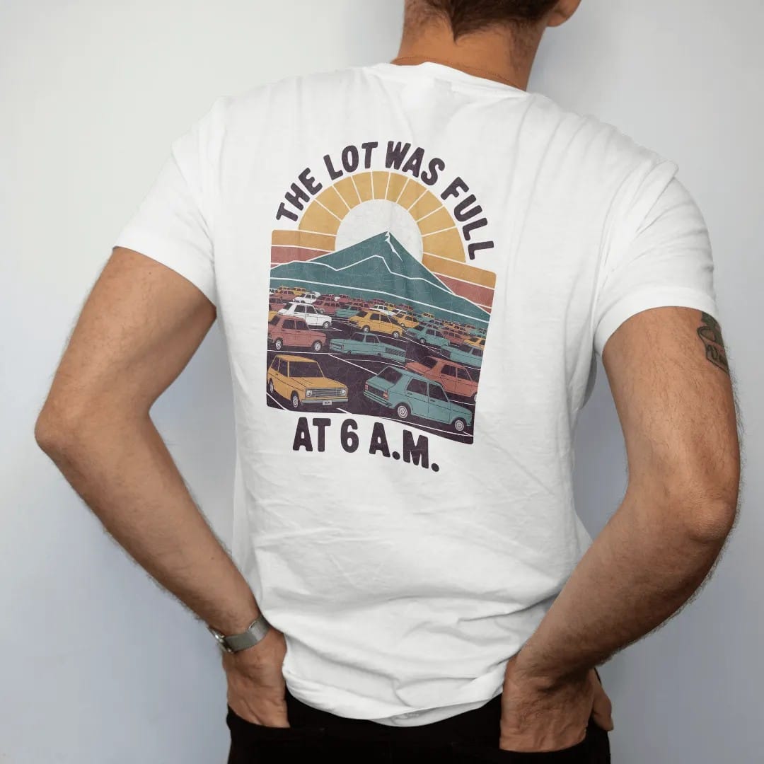 Man wearing a White tee, seen from the back: a packed trailhead parking lot with tiny cars and a mountain behind, a sad sun rising, faded 70s souvenir style, lettered "THE LOT WAS FULL / AT 6 A.M." in chunky rounded 70s letters with a drop shadow,…