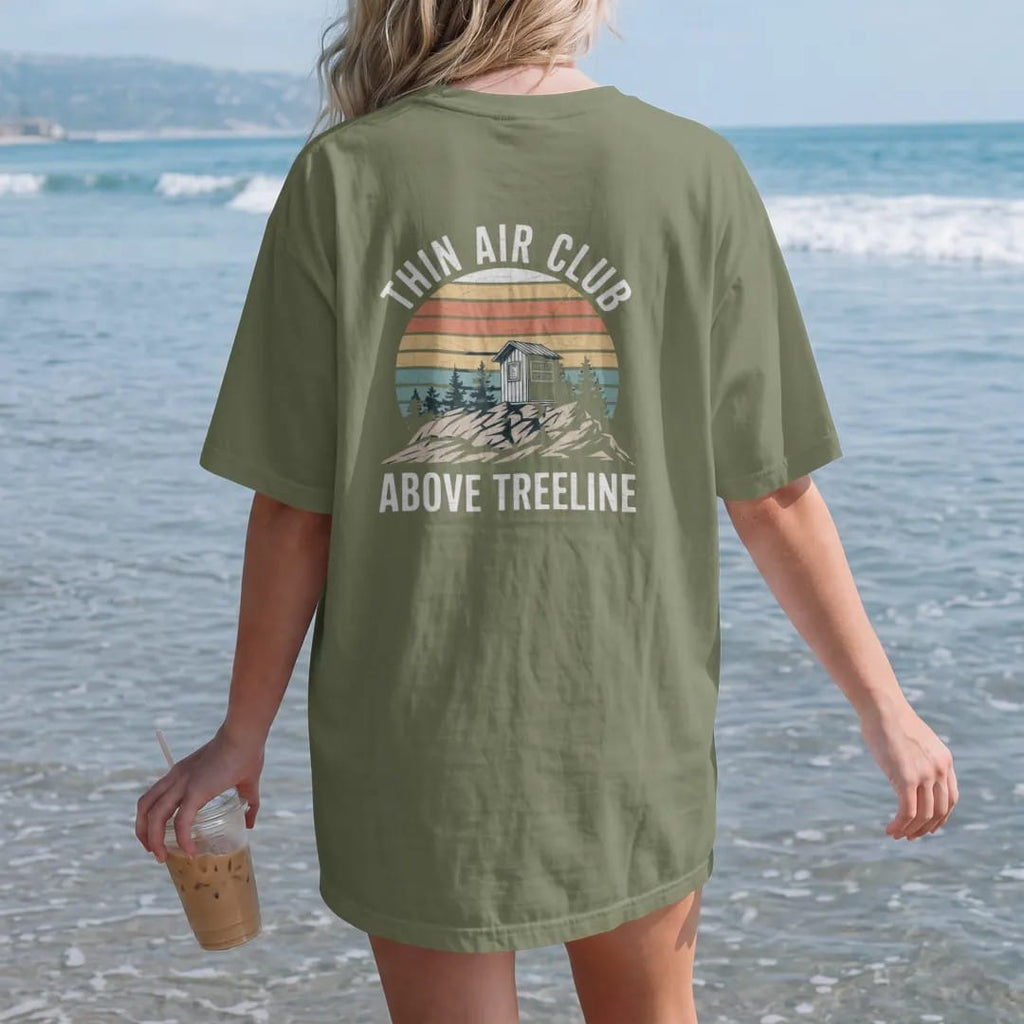 Woman on the beach in a Moss tee, back view: a rocky alpine summit above the last scrubby trees with a small summit register box, faded 70s souvenir style, lettered "THIN AIR CLUB / ABOVE TREELINE" in chunky rounded 70s letters with a drop shadow,…