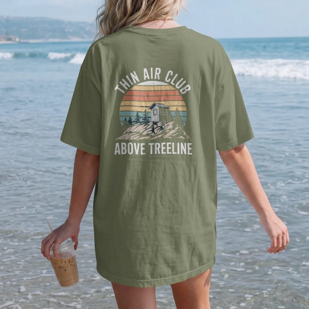 Woman on the beach in a Moss tee, back view: a rocky alpine summit above the last scrubby trees with a small summit register box, faded 70s souvenir style, lettered "THIN AIR CLUB / ABOVE TREELINE" in chunky rounded 70s letters with a drop shadow,…