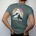 Man wearing a Blue Spruce tee, seen from the back: a long knife-edge ridge with a lone hiker walking it under huge clouds, wide panoramic layout, faded 70s souvenir style, lettered "RIDGELINE" in chunky rounded 70s letters with a drop shadow,…