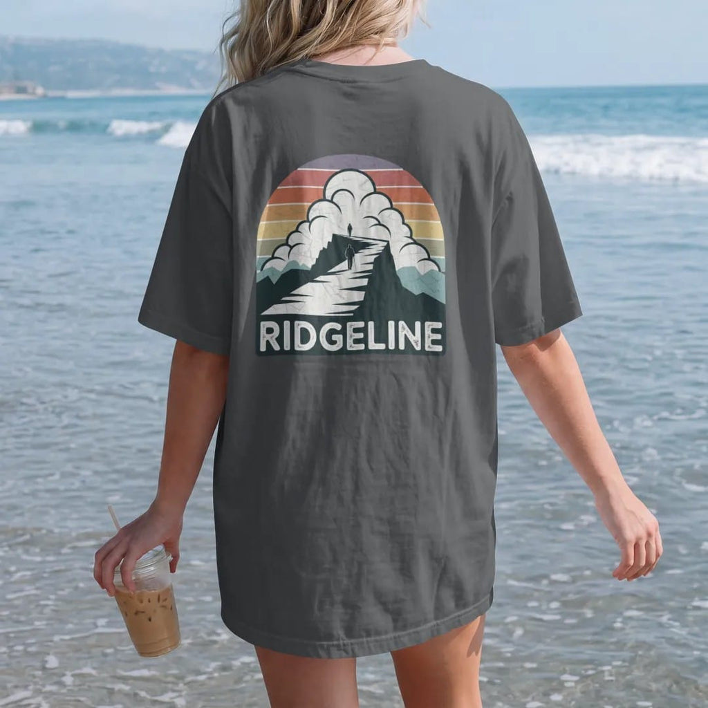 Woman on the beach in a Graphite tee, back view: a long knife-edge ridge with a lone hiker walking it under huge clouds, wide panoramic layout, faded 70s souvenir style, lettered "RIDGELINE" in chunky rounded 70s letters with a drop shadow, printed…
