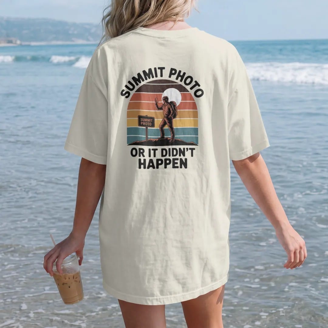 Woman on the beach in a Ivory tee, back view: a hiker posing with a phone beside a summit sign under a bright sun, faded 70s souvenir style, lettered "SUMMIT PHOTO / OR IT DIDN'T HAPPEN" in chunky rounded 70s letters with a drop shadow, printed in…