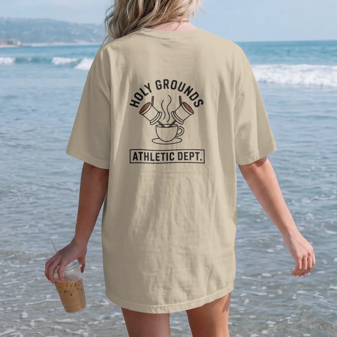 Woman on the beach in a Sandstone tee, back view: two crossed portafilters behind a steaming cup with Holy Grounds arched in spaced caps and Athletic Dept. underneath, drawn in a single fine crema-cream line with one burnt copper accent.