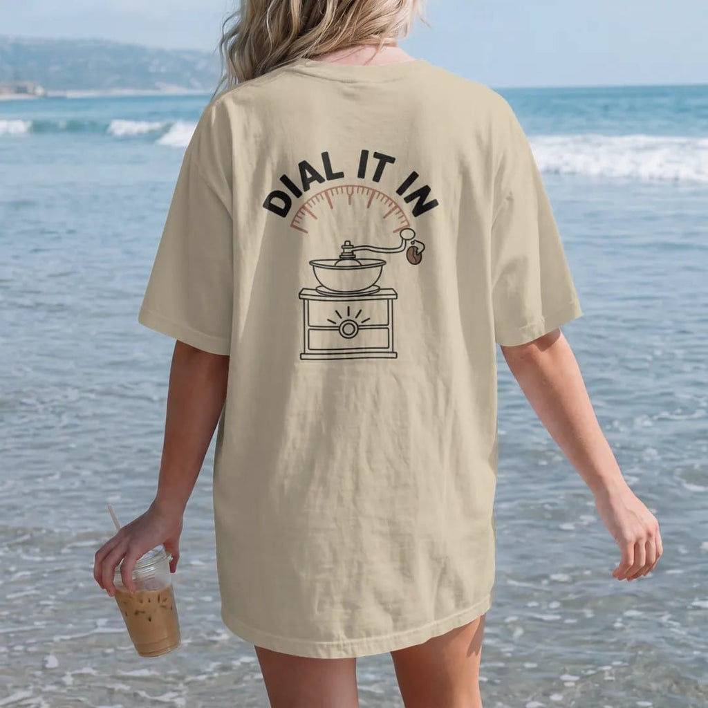 Woman on the beach in a Sandstone tee, back view: a coffee-bean mascot turning the dial of a burr grinder with Dial It In arched over it like a gauge, drawn in a single fine crema-cream line with one burnt copper accent.