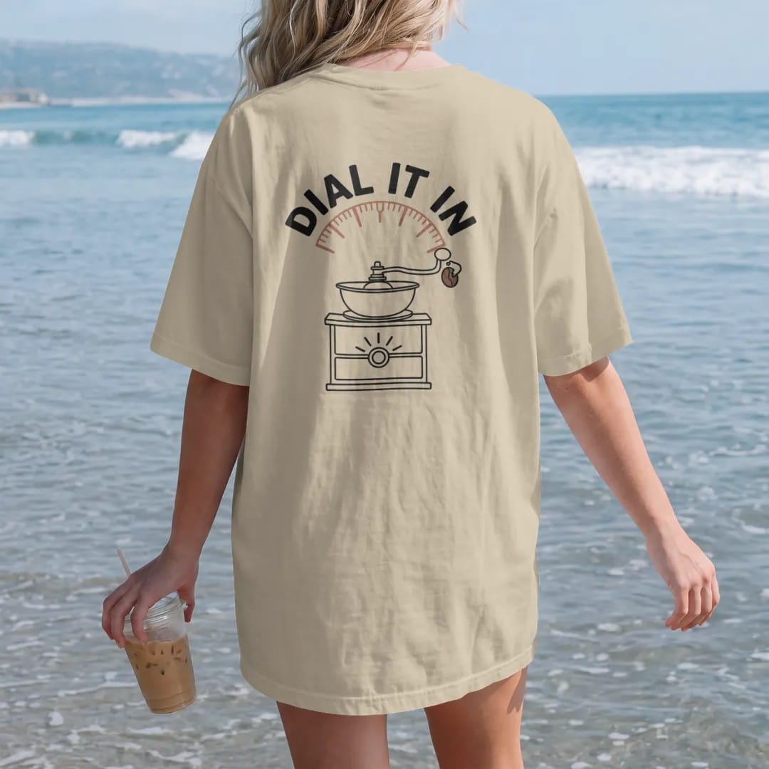 Woman on the beach in a Sandstone tee, back view: a coffee-bean mascot turning the dial of a burr grinder with Dial It In arched over it like a gauge, drawn in a single fine crema-cream line with one burnt copper accent.