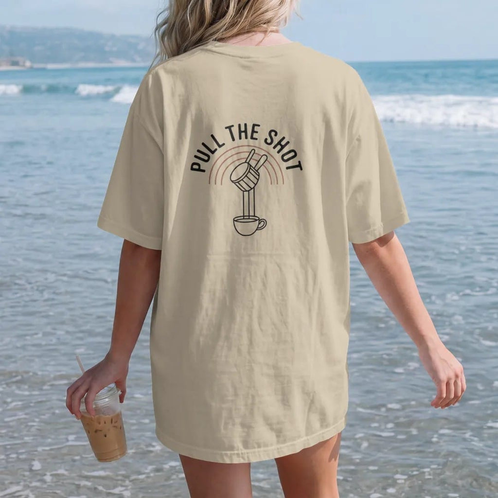 Woman on the beach in a Sandstone tee, back view: a portafilter dripping two streams of espresso into a cup over thin arc lines with Pull The Shot stacked on top, drawn in a single fine crema-cream line with one burnt copper accent.