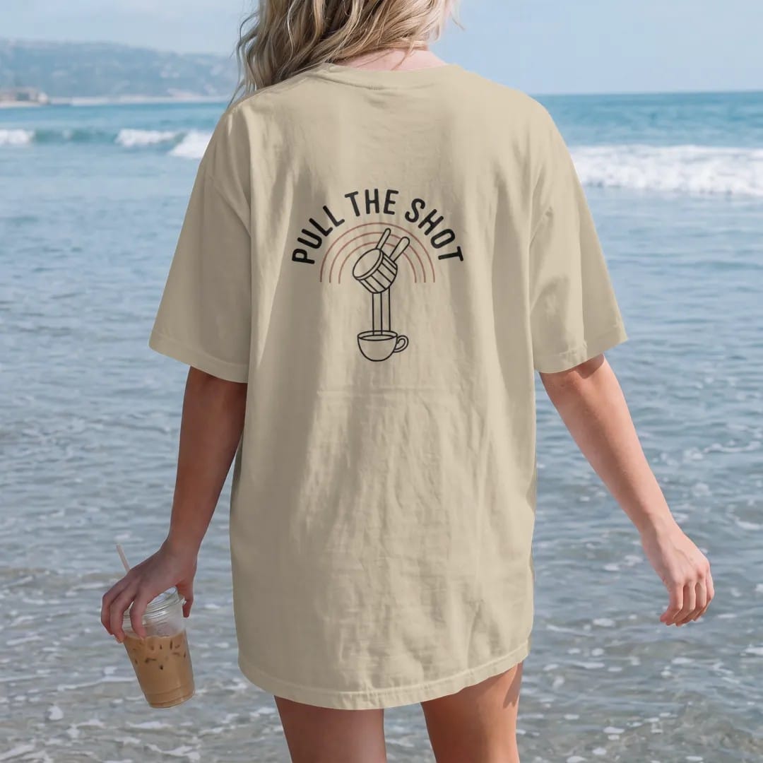 Woman on the beach in a Sandstone tee, back view: a portafilter dripping two streams of espresso into a cup over thin arc lines with Pull The Shot stacked on top, drawn in a single fine crema-cream line with one burnt copper accent.