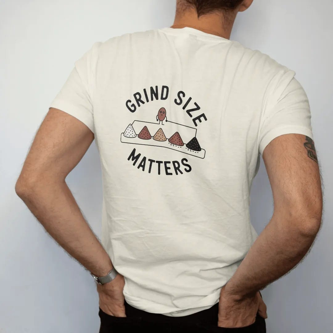 Man wearing a Ivory tee, seen from the back: a reference row of coffee piles from coarse to fine with a bean mascot pointing at them, under Grind Size and over Matters, drawn in a single fine crema-cream line with one burnt copper accent.