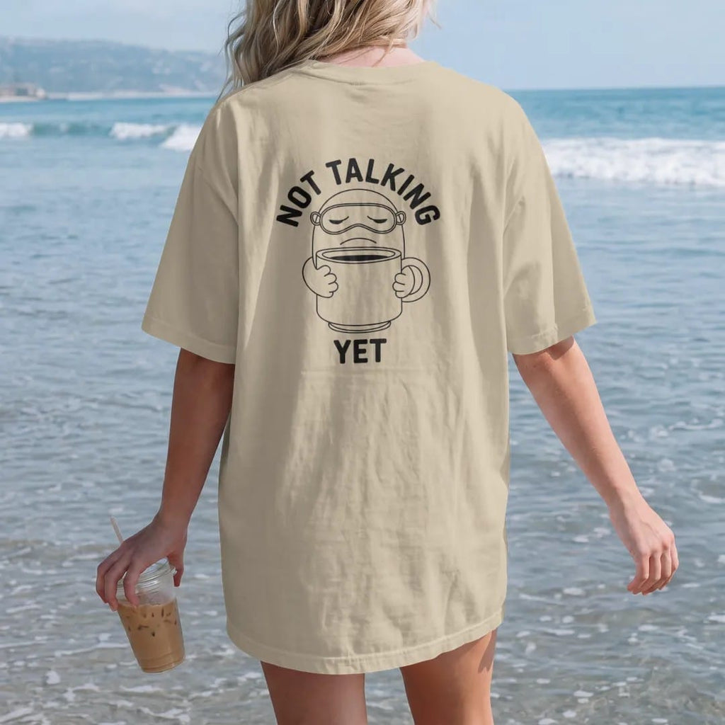 Woman on the beach in a Sandstone tee, back view: a grumpy coffee-bean mascot with a pushed-up sleep mask hugging an oversized mug, under Not Talking and over a small Yet, drawn in a single fine crema-cream line with one burnt copper accent.