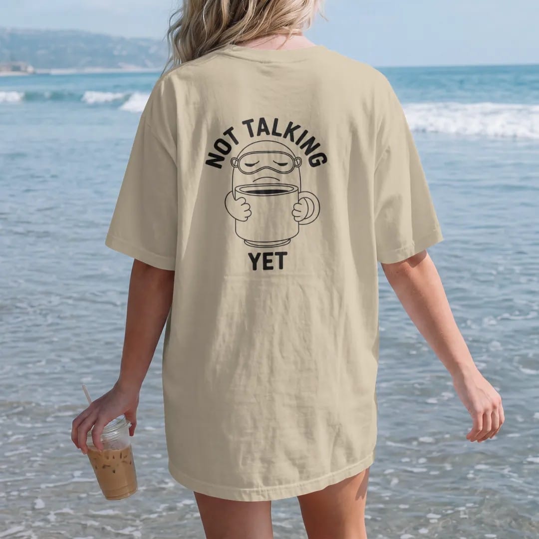 Woman on the beach in a Sandstone tee, back view: a grumpy coffee-bean mascot with a pushed-up sleep mask hugging an oversized mug, under Not Talking and over a small Yet, drawn in a single fine crema-cream line with one burnt copper accent.