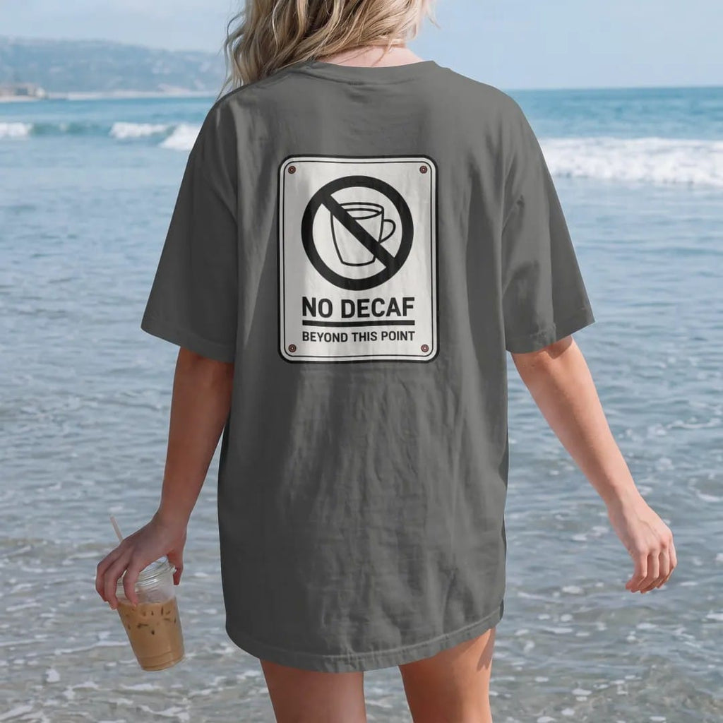 Woman on the beach in a Pepper tee, back view: a warning-sign parody with a no-symbol over a decaf cup, No Decaf in caps and Beyond This Point below, drawn in a single fine crema-cream line with one burnt copper accent.