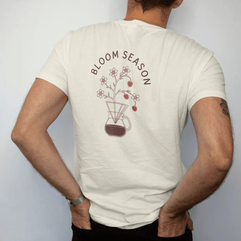 Man wearing a Ivory tee, seen from the back: a blossoming coffee branch with red cherries growing out of a pour-over cone under Bloom Season in spaced caps, drawn in a single fine roast-brown line with one burnt copper accent.