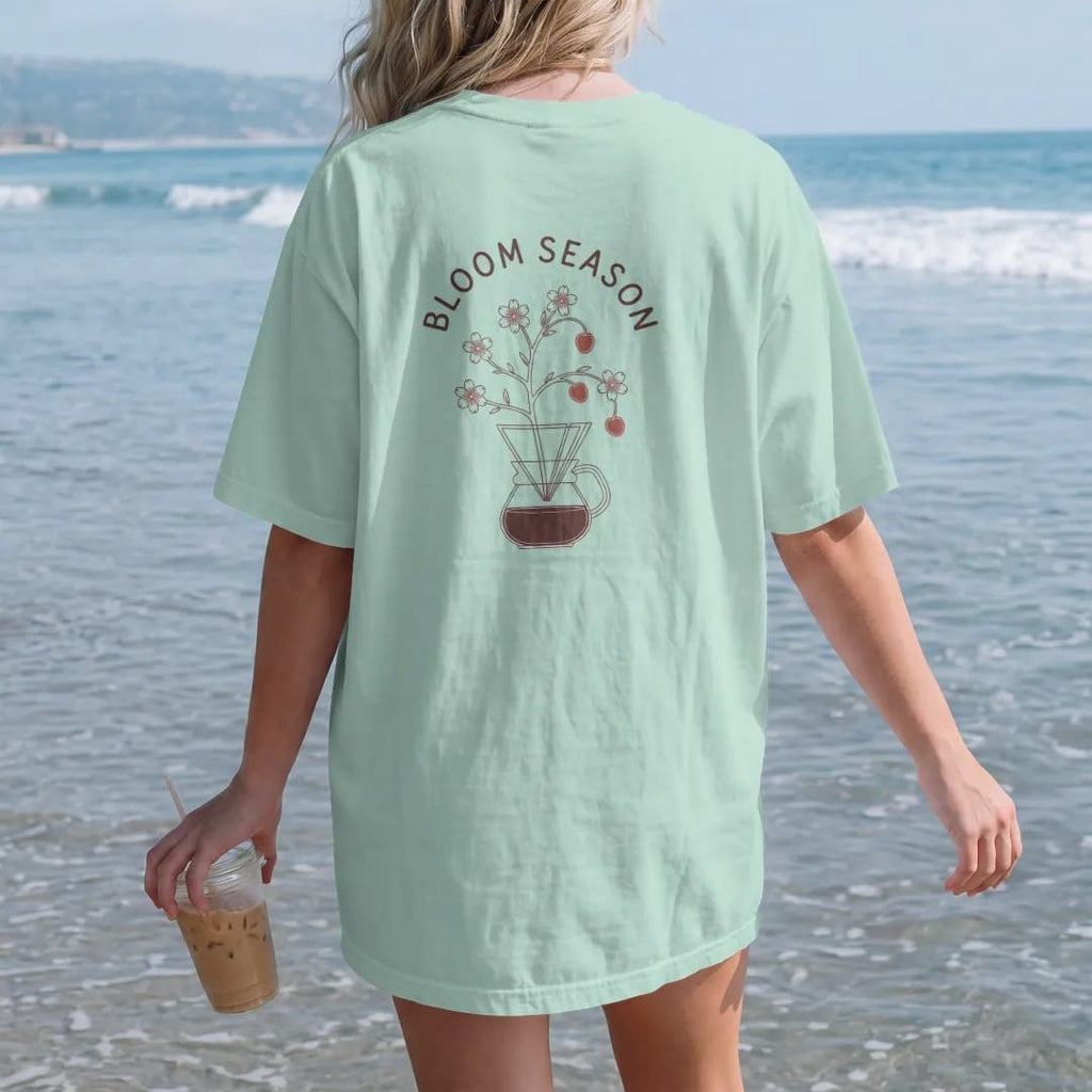 Woman on the beach in a Chalky Mint tee, back view: a blossoming coffee branch with red cherries growing out of a pour-over cone under Bloom Season in spaced caps, drawn in a single fine roast-brown line with one burnt copper accent.