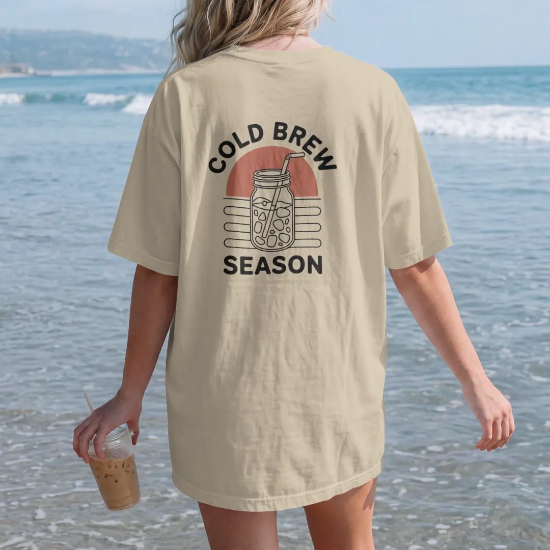 Woman on the beach in a Sandstone tee, back view: a mason jar of iced cold brew with a straw in front of a simple outlined sun, with Cold Brew curved above and Season below, drawn in a single fine crema-cream line with one burnt copper accent.