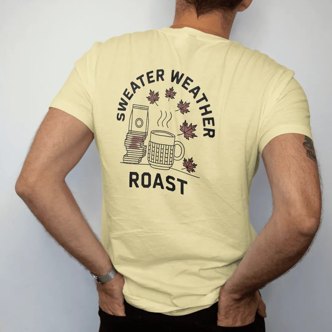Man wearing a Butter tee, seen from the back: a steaming mug in a knit sleeve next to coffee bags and falling maple leaves under Sweater Weather with Roast below, drawn in a single fine roast-brown line with one burnt copper accent.