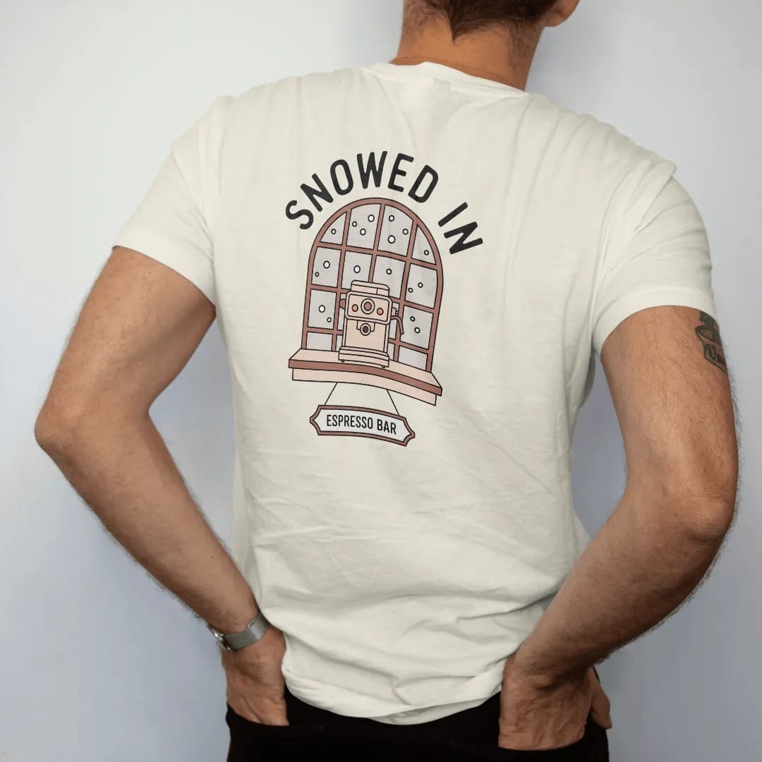 Man wearing a Ivory tee, seen from the back: a small espresso machine before a frosted snowy window under Snowed In with a hanging Espresso Bar sign, drawn in a single fine crema-cream line with one burnt copper accent.