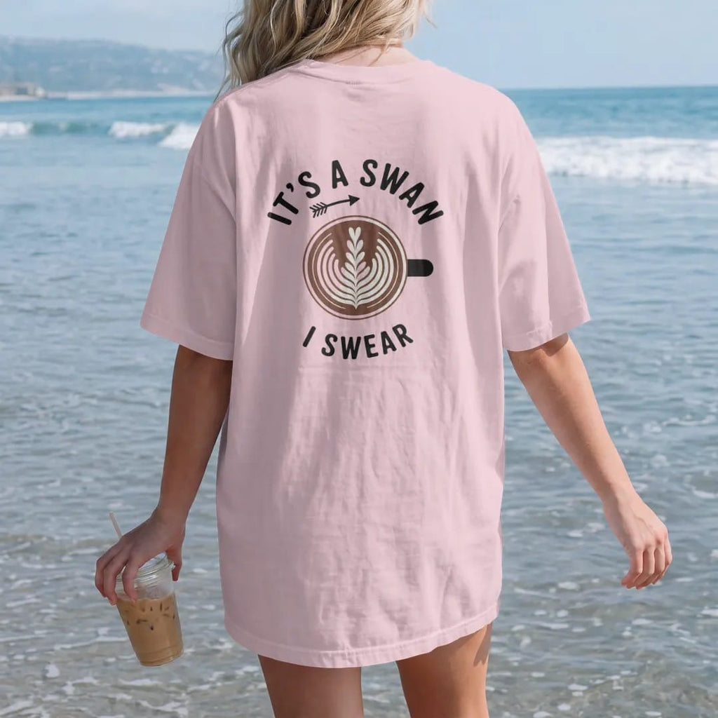 Woman on the beach in a Blossom tee, back view: a latte from above with a lopsided foam blob and an arrow, It's A Swan stacked above and a tiny I Swear below, drawn in a single fine roast-brown line with one burnt copper accent.