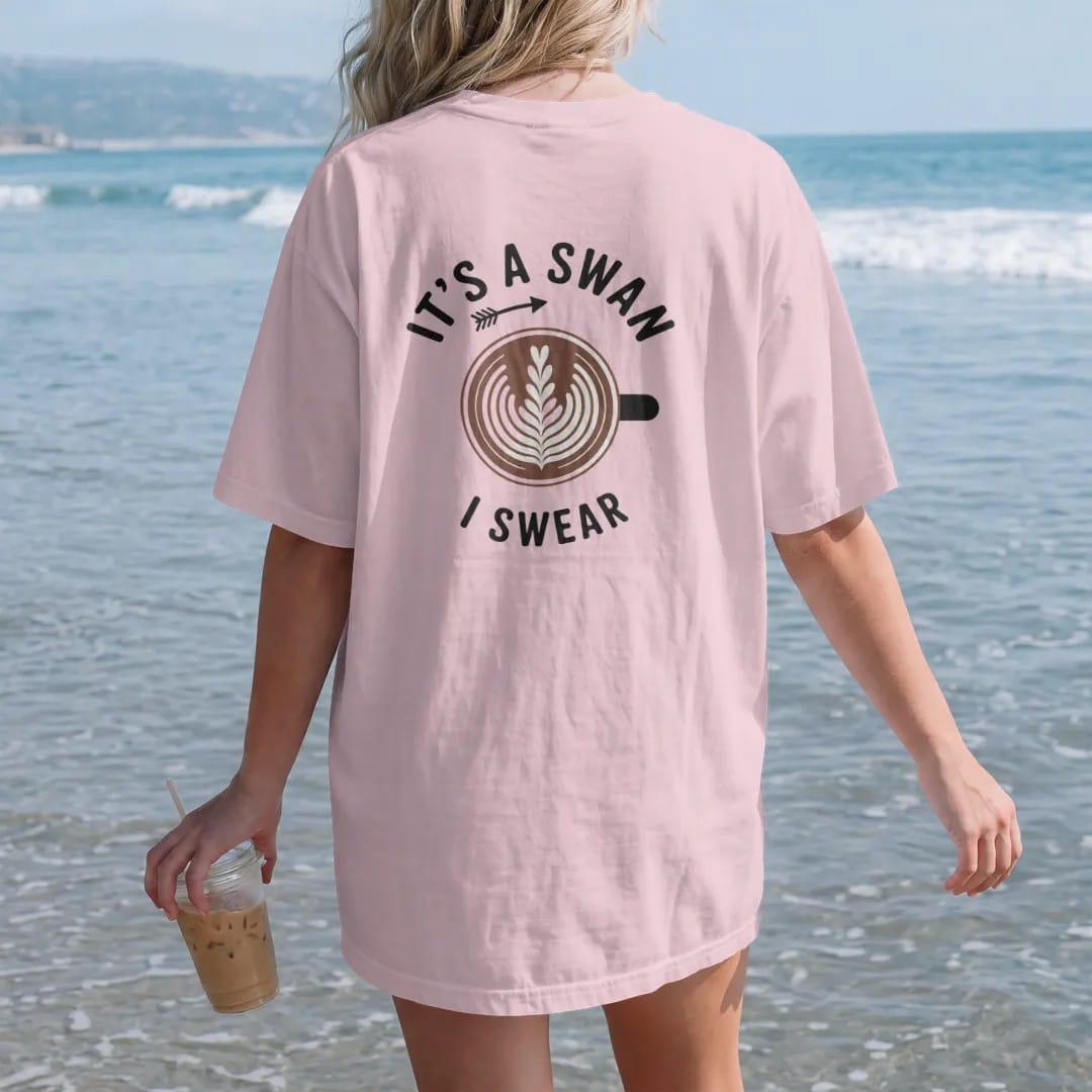 Woman on the beach in a Blossom tee, back view: a latte from above with a lopsided foam blob and an arrow, It's A Swan stacked above and a tiny I Swear below, drawn in a single fine roast-brown line with one burnt copper accent.