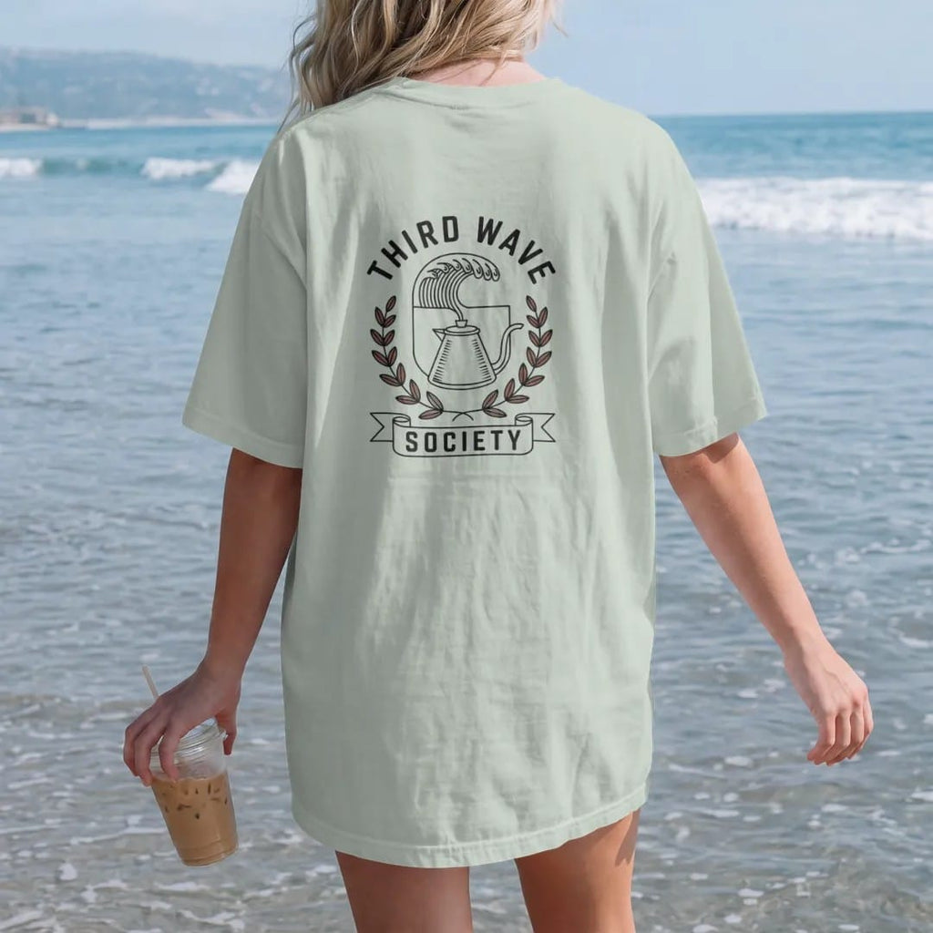 Woman on the beach in a Bay tee, back view: a crest of an ocean wave pouring from a gooseneck kettle in coffee-leaf laurels, Third Wave arched above and Society on a ribbon, drawn in a single fine roast-brown line with one burnt copper accent.
