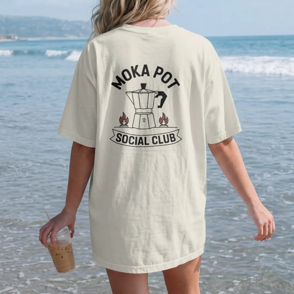Woman on the beach in a Ivory tee, back view: a sputtering stovetop moka pot over a small gas flame with Moka Pot curved above and Social Club in a banner, drawn in a single fine roast-brown line with one burnt copper accent.