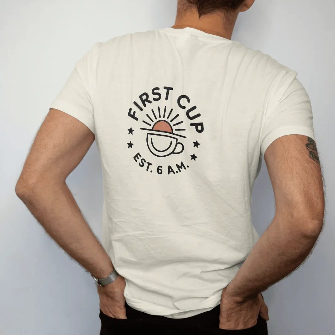 Man wearing a Ivory tee, seen from the back: a round badge of a sunrise rising over a coffee cup with First Cup curved on top and Est. 6 A.M. along the bottom, drawn in a single fine crema-cream line with one burnt copper accent.