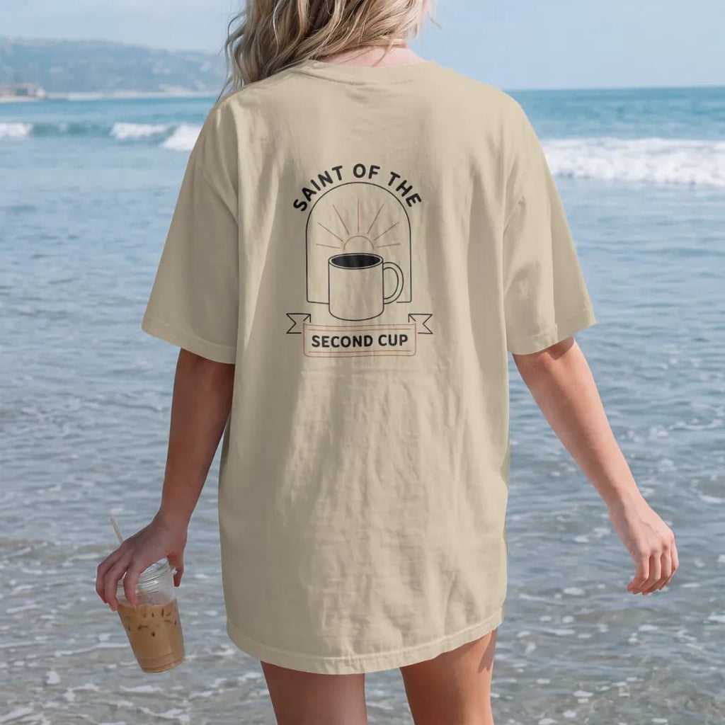 Woman on the beach in a Sandstone tee, back view: a haloed refilled mug in a thin arched window outline, Saint Of The arched above and Second Cup on a ribbon, drawn in a single fine crema-cream line with one burnt copper accent.