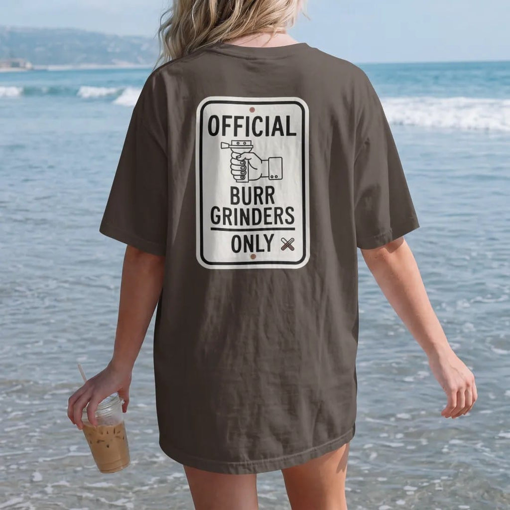 Woman on the beach in a Espresso tee, back view: a street-sign parody with a hand burr grinder icon reading Burr Grinders Only, with a crossed-out blade grinder in the corner, drawn in a single fine crema-cream line with one burnt copper accent.