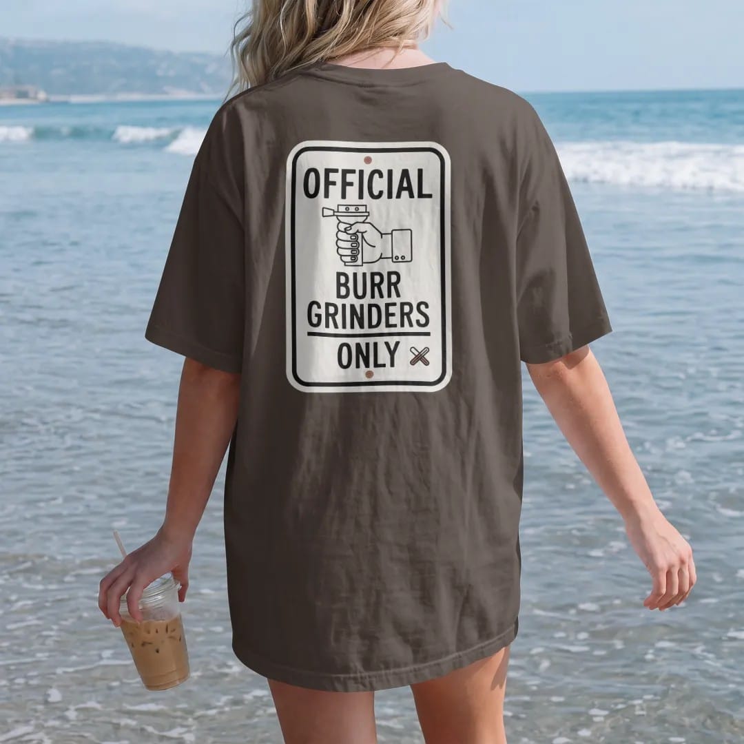 Woman on the beach in a Espresso tee, back view: a street-sign parody with a hand burr grinder icon reading Burr Grinders Only, with a crossed-out blade grinder in the corner, drawn in a single fine crema-cream line with one burnt copper accent.