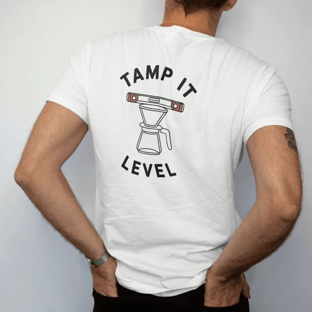 Man wearing a White tee, seen from the back: a tamper pressing into a portafilter with a bubble level balanced on top, Tamp It above and Level below, drawn in a single fine crema-cream line with one burnt copper accent.