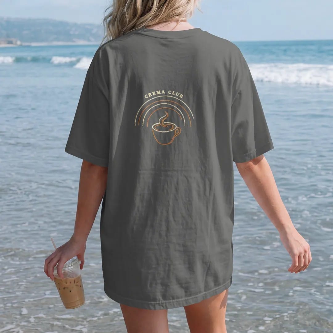 Woman on the beach in a Pepper tee, back view: an espresso cup with a thick crema swirl in front of thin arc lines under an arched Crema Club header, drawn in a single fine crema-cream line with one burnt copper accent.