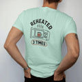 Man wearing a Chalky Mint tee, seen from the back: an open microwave with a sad half-full mug inside, Reheated across the top and a 3 Times stamp below, drawn in a single fine roast-brown line with one burnt copper accent.