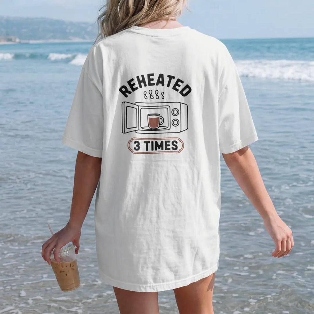 Woman on the beach in a White tee, back view: an open microwave with a sad half-full mug inside, Reheated across the top and a 3 Times stamp below, drawn in a single fine roast-brown line with one burnt copper accent.