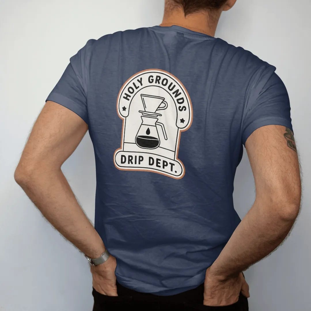 Man wearing a Navy tee, seen from the back: a department-style badge with a pour-over dripper and carafe, Holy Grounds curved on top and Drip Dept. in the center bar, drawn in a single fine crema-cream line with one burnt copper accent.