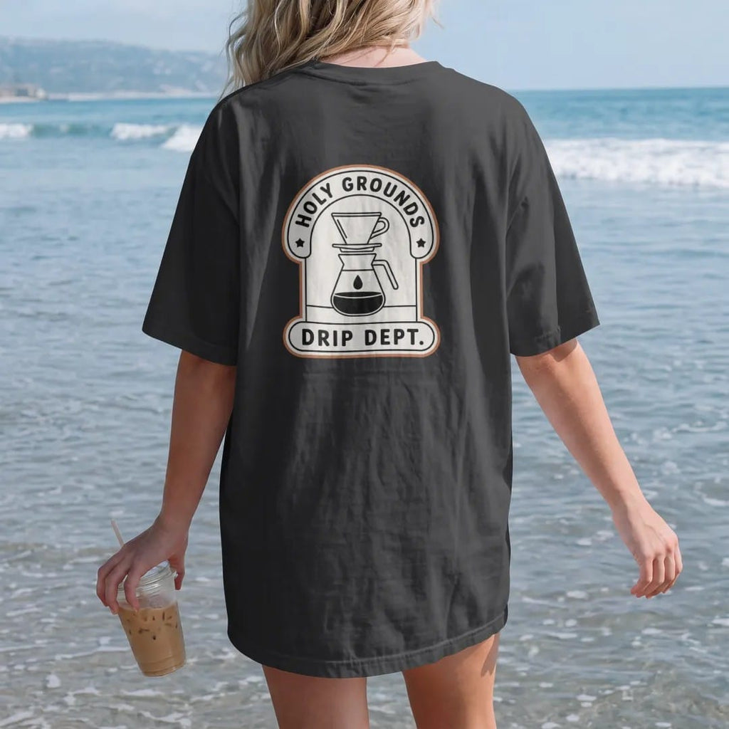 Woman on the beach in a Black tee, back view: a department-style badge with a pour-over dripper and carafe, Holy Grounds curved on top and Drip Dept. in the center bar, drawn in a single fine crema-cream line with one burnt copper accent.