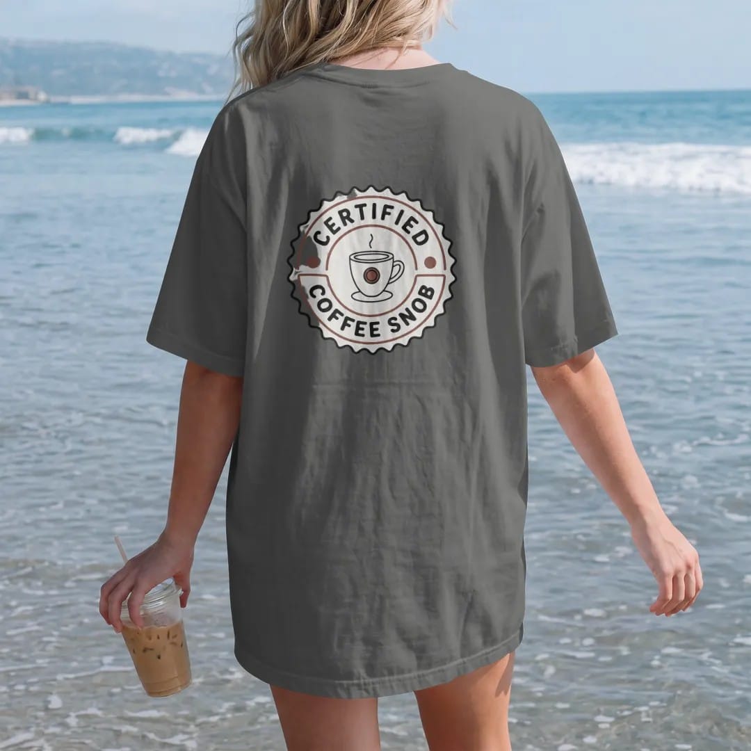 Woman on the beach in a Pepper tee, back view: a scalloped official seal with a monocled coffee cup, Certified across the top and Coffee Snob on a ribbon, drawn in a single fine roast-brown line with one burnt copper accent.
