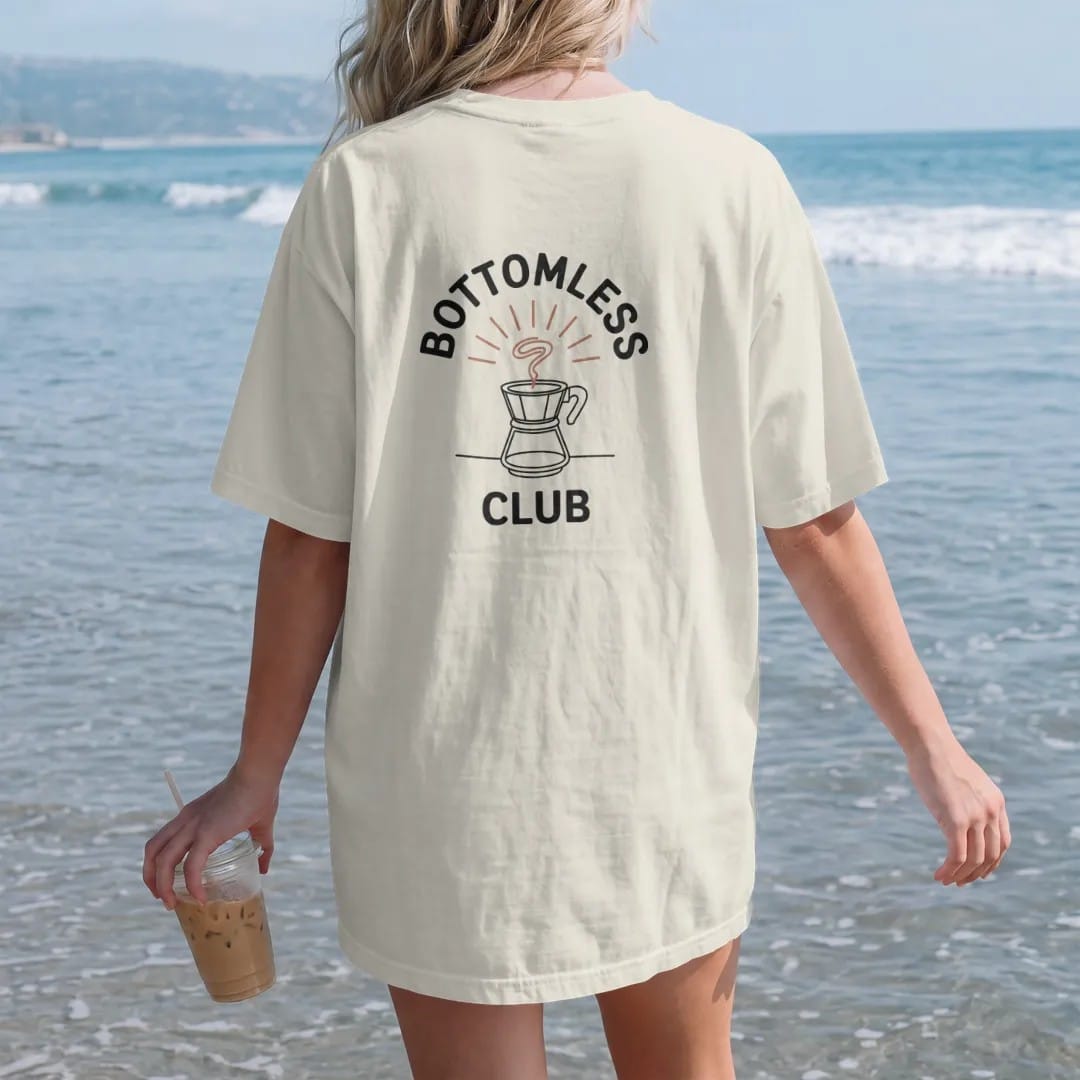 Woman on the beach in a Ivory tee, back view: a bottomless portafilter seen from below with a spiraling espresso stream over a few thin rays, Bottomless above and Club below, drawn in a single fine crema-cream line with one burnt copper accent.