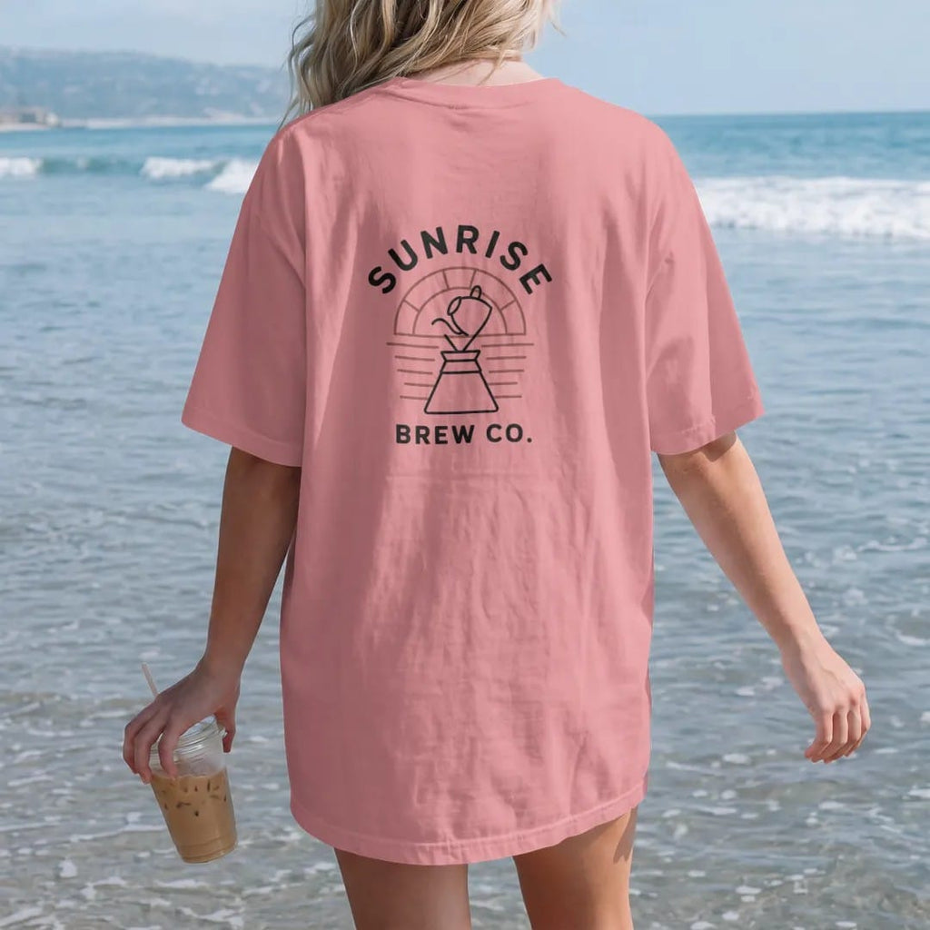 Woman on the beach in a Watermelon tee, back view: a kettle pouring into a pour-over cone in front of a simple outlined rising sun, with Sunrise arched above and Brew Co. below, drawn in a single fine roast-brown line with one burnt copper accent.