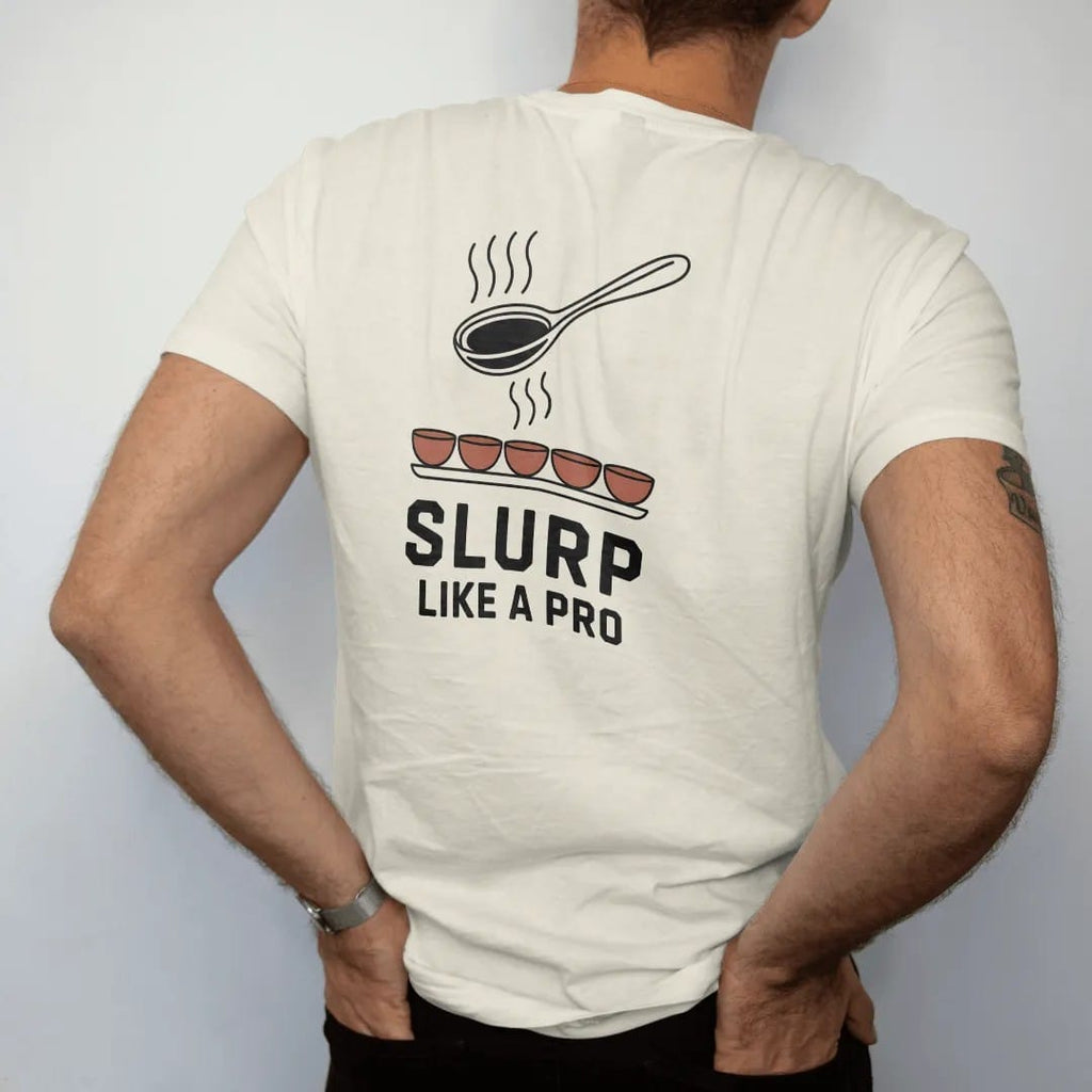 Man wearing a Ivory tee, seen from the back: a cupping spoon at a cartoon mouth mid-slurp over a row of cupping bowls, with Slurp in caps and Like A Pro below, drawn in a single fine crema-cream line with one burnt copper accent.